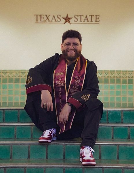 Happy graduate in maroon stole and cap sits on green tiled steps below the 'Texas State' sign