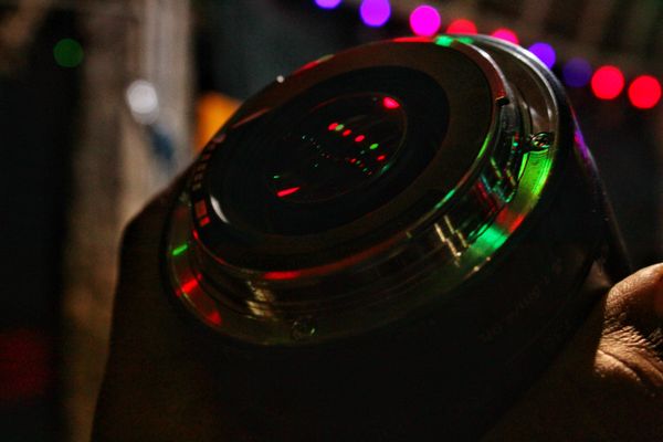 Close-up of a camera lens held by a hand, reflecting red and green festive lights with blurred bokeh background