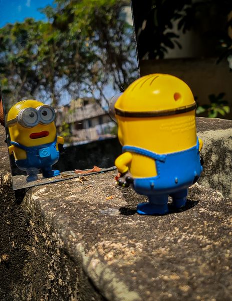 A yellow Minion toy looks into a mirror reflecting green trees and a distant building from above a stone wall