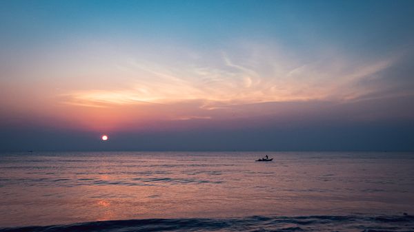 Dramatic sunset over a calm ocean, casting pink and blue hues across the sky with a small boat
