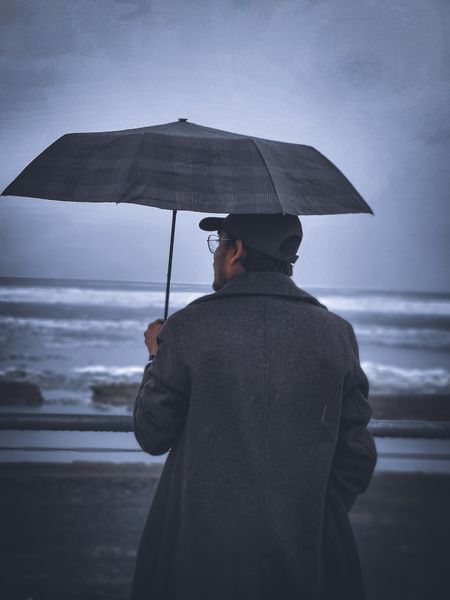 A person in a coat holds an umbrella, looking out at a stormy ocean under a grey sky