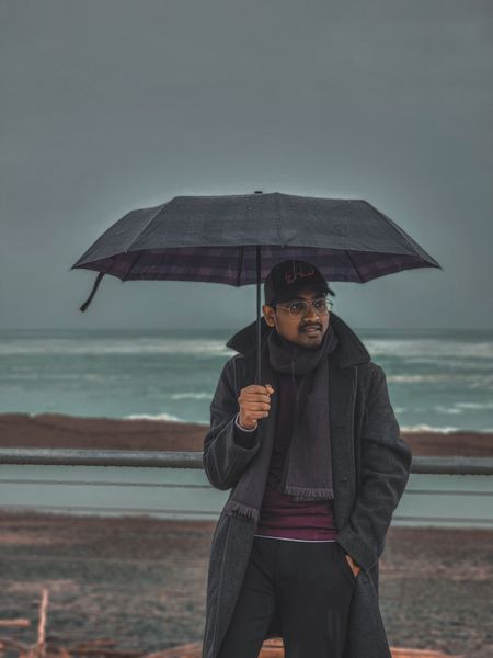 Man in a coat and hat holds an umbrella, looking thoughtfully at the stormy sea under a grey sky