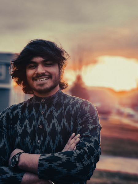 Smiling young man with long dark hair and a patterned shirt, arms crossed, bathed in warm sunset light