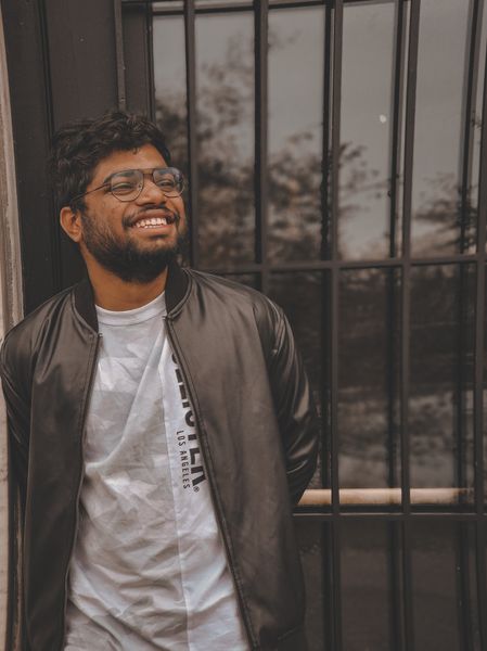 Smiling young man with glasses and a beard in a black jacket leans against a window grill