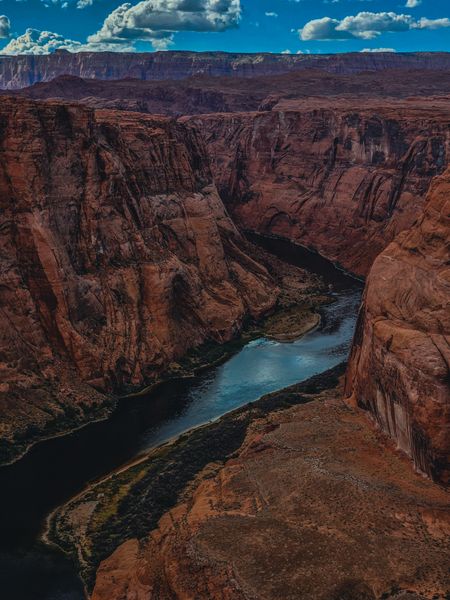 A deep canyon with towering red rock walls, a winding blue-green river below, under a cloudy blue sky