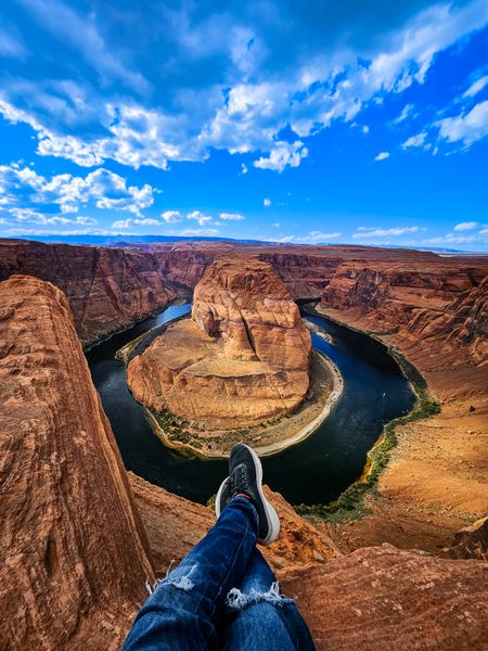 Person's legs dangle above Horseshoe Bend, showing the Colorado River snaking through a red rock canyon under blue sky