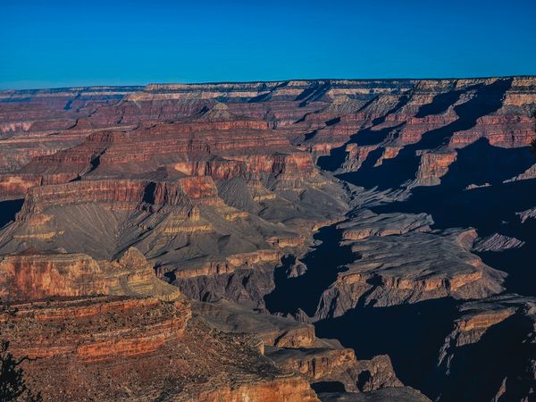 Vast panorama of the Grand Canyon with deep orange and red rock layers, contrasting shadows, and a bright blue sky