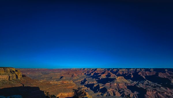 A panoramic view of the vast Grand Canyon under a clear, deep blue sky, with rugged rock formations and dramatic shadows