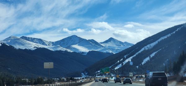 Cars driving on a highway through a snowy mountain valley with a vibrant blue sky and white clouds