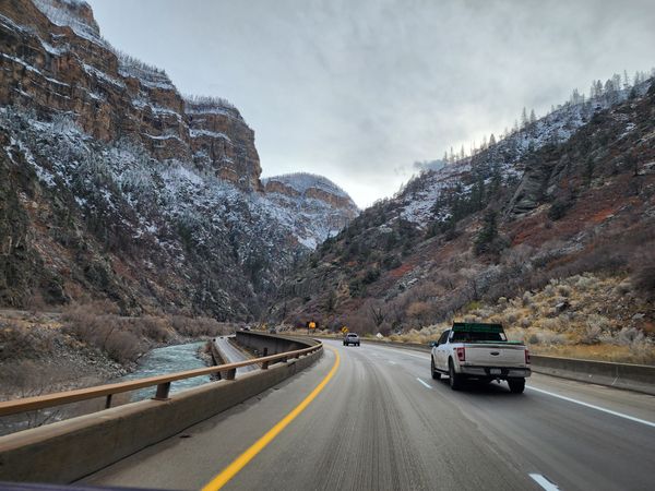 Vehicles drive on a winding highway through a snowy mountain canyon alongside a turbulent river