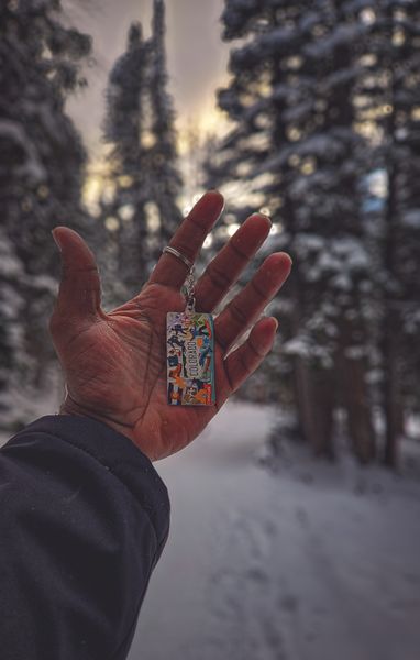 A person's hand holds a colorful 'Colorado' keychain against a blurred snowy forest background