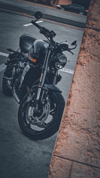 A black Triumph motorcycle with orange accents parked on asphalt next to a textured stone wall, car in background