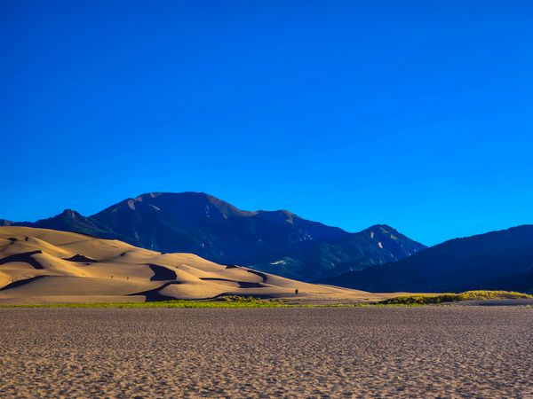 Expansive golden sand dunes ripple towards dark, majestic mountains under a clear, deep blue sky