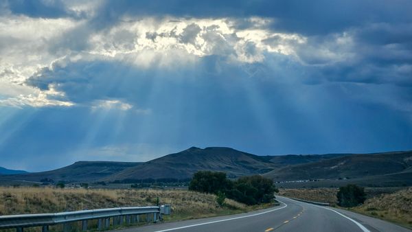 A scenic road winding through dry fields towards distant mountains under a dramatic sky with sun rays breaking through clouds