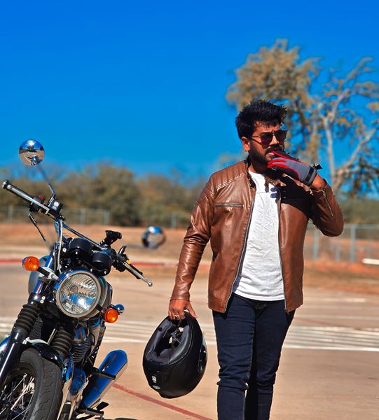 Stylish man in a brown leather jacket and sunglasses holding a helmet, standing next to a motorcycle under a clear blue sky