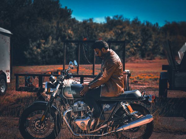 A man in a brown leather jacket sits on a Royal Enfield motorcycle in a rustic outdoor setting, looking down