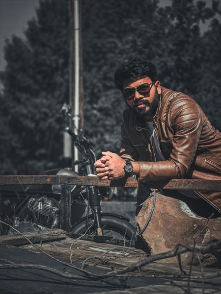 A stylish man in a brown leather jacket and sunglasses leans on a rustic structure with a motorcycle in the background