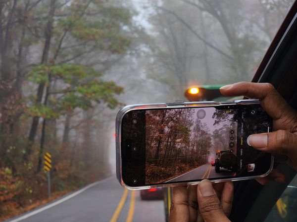 Hands hold a smartphone out a car window, capturing a foggy road winding through autumn trees
