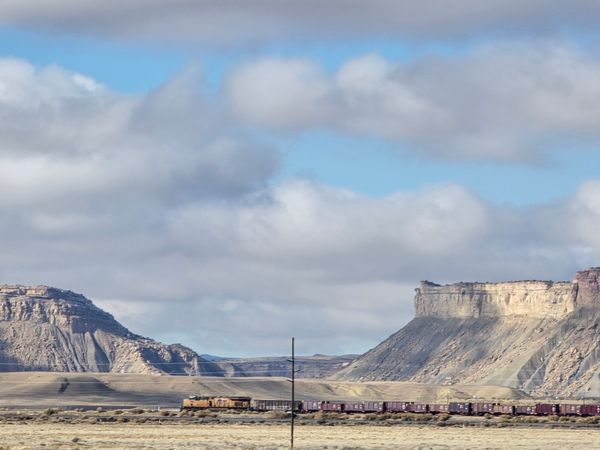 A long freight train moves through an arid, expansive landscape with flat-topped mesas under a cloudy sky