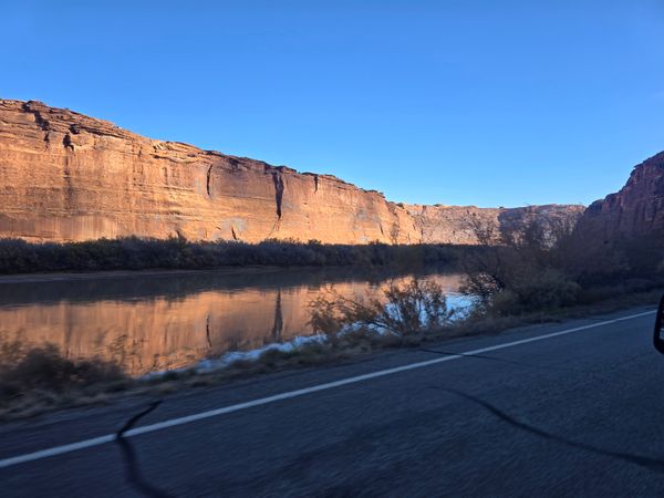 Scenic view of a wide river reflecting towering red sandstone cliffs under a clear blue sky, with a road in the foreground