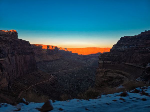 Dramatic canyon landscape during sunrise, glowing orange cliffs meet dark shadowed depths and a clear blue sky