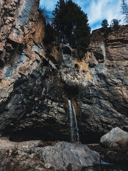 A narrow waterfall cascades down a massive, textured rock cliff, surrounded by trees under a blue sky