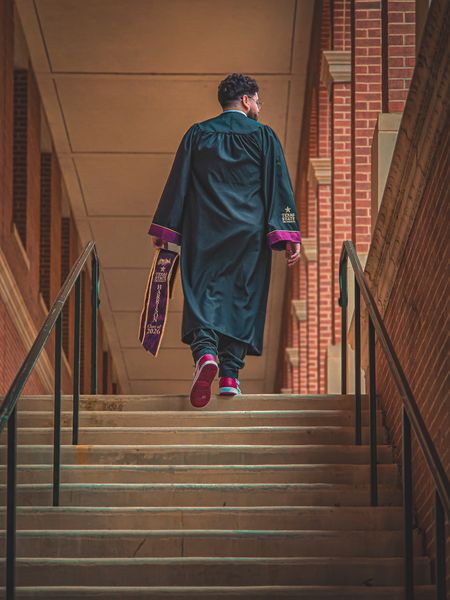 A graduate in a dark gown and red sneakers walks up brick stairs, holding a purple stole