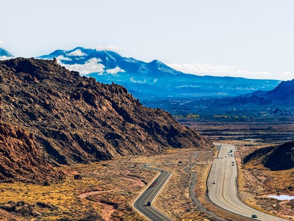 Highway winding through arid, rocky terrain with distant snow-capped mountains under a clear sky