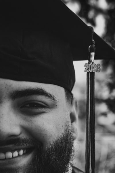 Close-up black and white portrait of a smiling graduate with a beard, wearing a cap with a '2026' tassel