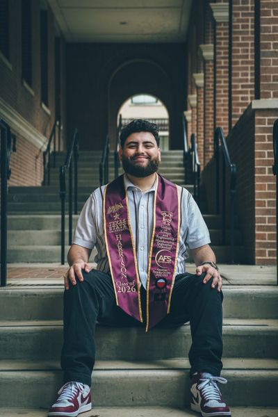 A smiling young man with a beard, wearing a maroon graduation stole, sits on stone steps outside a brick building