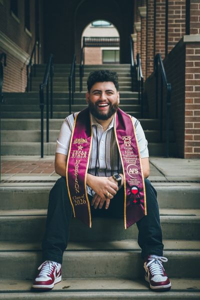 A smiling young man with a beard and maroon graduation stole sits on stone stairs in front of a brick building