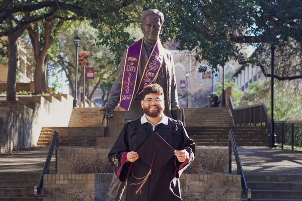 Smiling graduate in cap and gown holds a cap before a statue with a sash on a sunny university campus