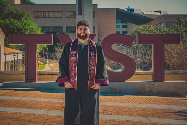 A smiling graduate in a black gown and maroon stole stands before large TXST letters on a university campus