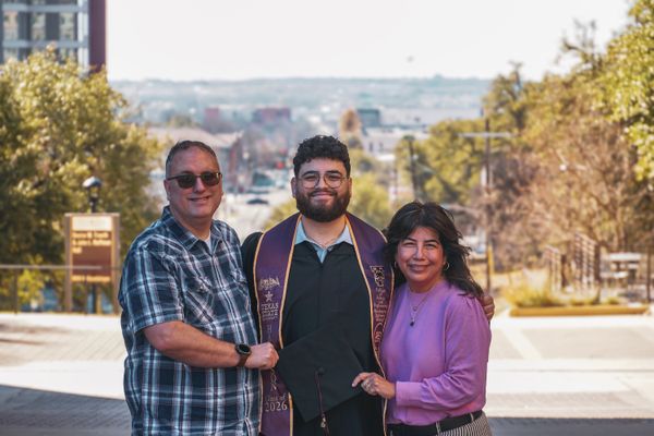 A smiling graduate in a purple stole stands between his proud parents on a sunny university campus