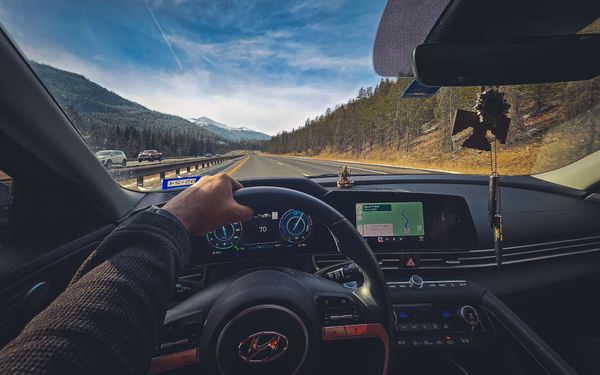 First-person view of driving a car on a highway through scenic mountains, showing steering wheel and dashboard with navigation