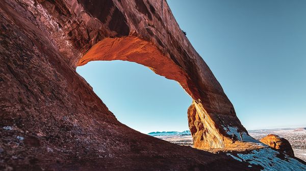 A majestic natural red rock arch frames a bright blue sky over a vast desert landscape with distant snowy mountains