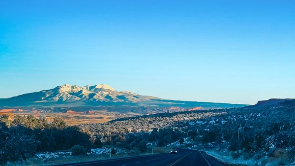 A winding highway leads towards majestic snow-capped mountains under a vast blue sky, with frosty trees and a distant car