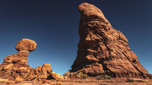 A dramatic desert landscape featuring a towering red rock formation and a unique balanced rock under a clear blue sky