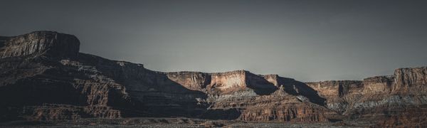 Panoramic view of rugged, layered canyons and mesas under a muted sky, with sunlight illuminating distant cliffs