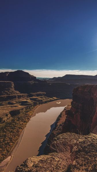 A wide, muddy river winds through a dramatic canyon under a vast blue sky, surrounded by rugged cliffs