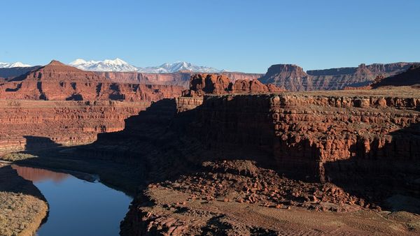 Red rock canyon with a winding river below, shadowed cliffs, and distant snow-capped mountains under a clear blue sky