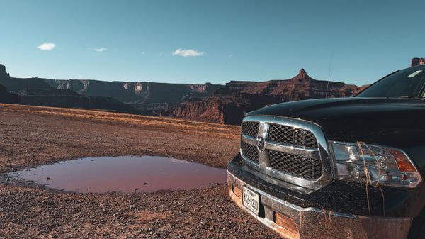 Black pickup truck parked on a dirt road by a red puddle, overlooking a grand desert canyon landscape