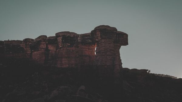 Sunlight streams through a dramatic natural arch in a dark, rugged rock formation against a muted sky