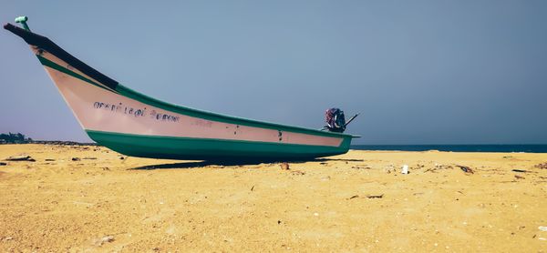 A traditional teal and white fishing boat with an outboard motor rests on a golden sandy beach, under a blue sky