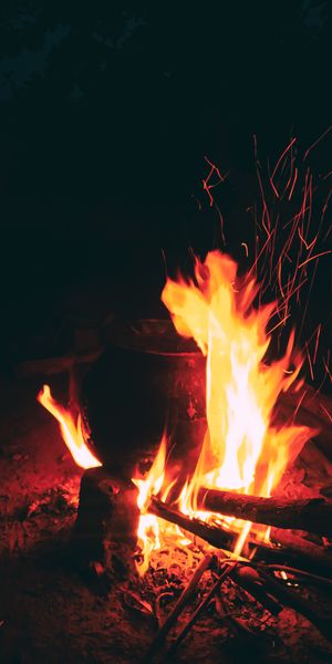 A vibrant campfire burning brightly at night, with sparks flying and a cooking pot over the flames