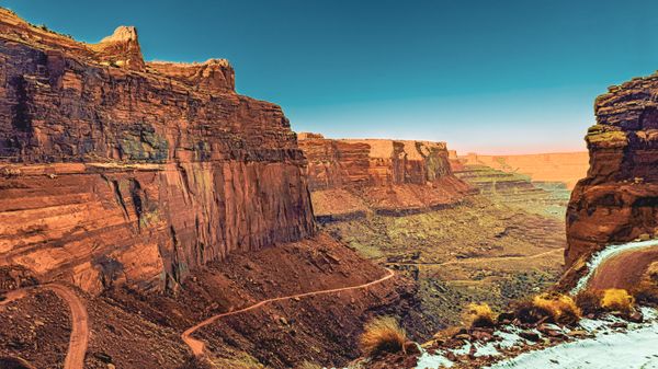 Vast canyon landscape with striking red rock formations, a winding dirt road, and a clear blue sky